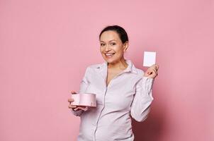 Pregnant middle-aged pretty woman smiles at camera while showing greeting card and gift box, isolated on pink background photo