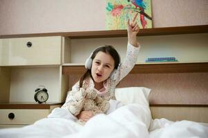 Adorable child girl waking up in happy mood, holding a plush toy and enjoying soundtrack on headphones, sitting on bed photo