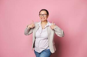 Happy middle-aged pregnant woman gestures with thumbs up, smiling cutely looking at camera over isolated pink background photo