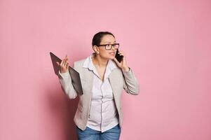 Multitasking overworked busy pregnant woman holding laptop and arguing while talking on mobile phone on pink backdrop photo