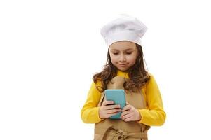 encantador niño chica, pequeño panadero confitero en del chef sombrero y beige delantal, participación teléfono inteligente, aislado blanco antecedentes foto