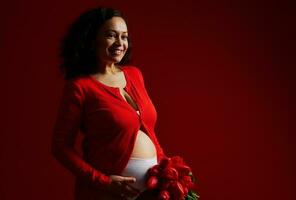 Beautiful happy pregnant woman caressing her belly, smiling looking away, posing with a bouquet of tulips, red backdrop photo