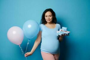 Beautiful pregnant brunette woman posing with newborn clothes and pink blue air balloons, isolated on blue background photo