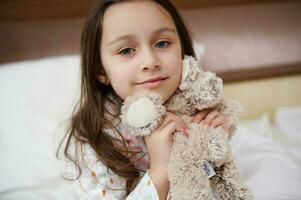 de cerca encantador pequeño niña en pijama, suavemente abrazando su felpa juguete y sonriente a cámara mientras despertar en el Mañana foto