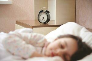 Focus on black alarm clock showing half past six, on bedside nightstand, in blurred foreground of a sleeping child girl photo