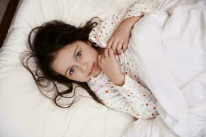 View from above of adorable little girl in pajamas, wrapped in white cozy duvet, lying on bed, looking at camera photo