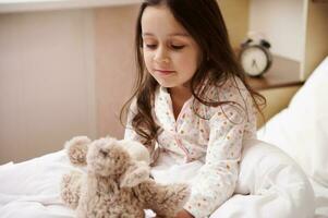 adorable niño niña en pijama, sentado en cama después despertar arriba en mañana, jugando con un felpa juguete en su acogedor dormitorio foto