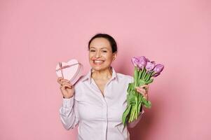 positivo mujer con ramo de flores de púrpura tulipanes y regalo caja, sonriente un con dientes sonrisa, mirando a cámara, aislado en rosado foto