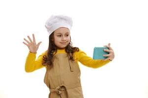 linda pequeño niña vistiendo blanco del chef sombrero y cocina delantal, taling en móvil teléfono vía vídeo enlace, aislado antecedentes foto