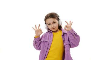 Lovely little school girl enjoys listening music on wireless headphones, shows OK sign, smiles cutely, looking at camera photo