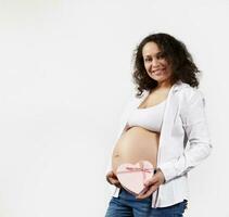 Smiling curly haired pregnant woman holding pink heart shaped gift box near her belly, isolated on white background photo