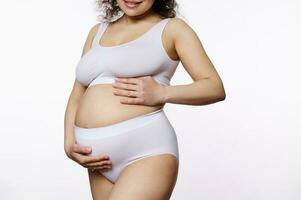 sección en medio embarazada mujer, expectante madre poniendo manos en su barriga, posando en blanco ropa interior en aislado antecedentes foto
