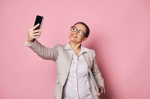 Attractive pregnant woman, business person wearing casual clothes and trendy glasses, carrying laptop and taking selfie photo