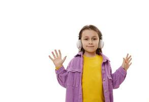 contento sonriente adorable pequeño niña en inalámbrico auriculares, mira y cámara y muestra su manos palmas arriba, blanco fondo foto