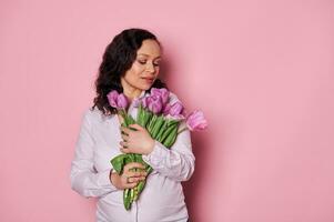 Delightful brunette, pregnant woman holding bunch of tulips on isolated pink background. Spring and femininity concept photo