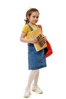 Smart schoolgirl in yellow t-shirt and denim sundress, carrying backpack and school supplies, smiles on white background photo