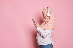 Side portrait of happy Arab Muslim woman in pink hijab with headphones and mobile phone, isolated on color background photo