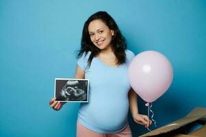 Ethnic pregnant woman with ultrasound scan of her newborn baby, smiles at camera, holds a pink balloon, isolated on blue photo