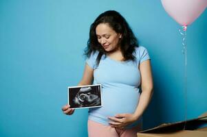 Happy caring loving expectant mother, pregnant woman touching belly, holding ultrasound image of baby on blue background photo