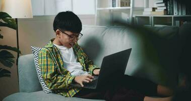 ordinateur de poche tir, asiatique garçon portant des lunettes écolier en utilisant une portable ordinateur tandis que séance sur le canapé à maison. éducation, e-learning et mode de vie concepts. video