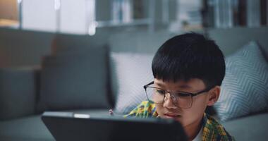 palmare sparo, asiatico ragazzo indossare bicchieri scolaro apprendimento in linea con un' digitale tavoletta e scrittura su fogli di lavoro a casa. formazione scolastica, e-learning e stile di vita concetti. video