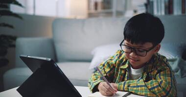 palmare sparo, asiatico ragazzo indossare bicchieri scolaro apprendimento in linea con un' digitale tavoletta e scrittura su fogli di lavoro a casa. formazione scolastica, e-learning e stile di vita concetti. video