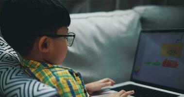 muñequita disparo, asiático chico vistiendo lentes colegial utilizando un ordenador portátil computadora mientras sentado en el sofá a hogar. educación, e-learning y estilo de vida conceptos. video