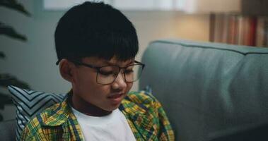 chariot tir, asiatique garçon portant des lunettes écolier en utilisant une portable ordinateur tandis que séance sur le canapé à maison. éducation, e-learning et mode de vie concepts. video