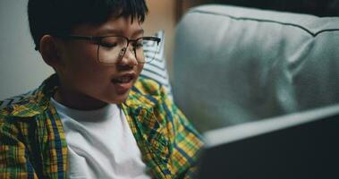 lado vista, asiático chico vistiendo lentes colegial utilizando un ordenador portátil computadora mientras sentado en el sofá a hogar. educación, e-learning y estilo de vida conceptos. video