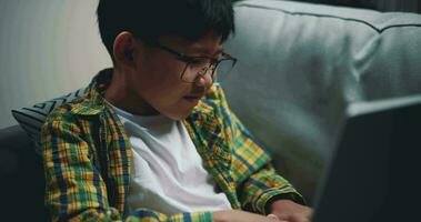 Mano disparo, asiático chico vistiendo lentes colegial utilizando un ordenador portátil computadora mientras sentado en el sofá a hogar. educación, e-learning y estilo de vida conceptos. video