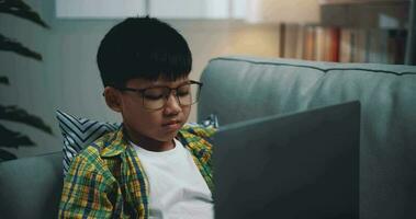 chariot tir, asiatique garçon portant des lunettes écolier en utilisant une portable ordinateur tandis que séance sur le canapé à maison. éducation, e-learning et mode de vie concepts. video
