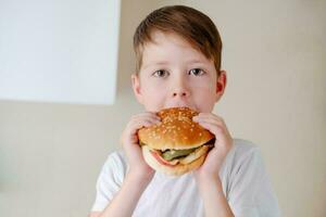 de cerca retrato de un pequeño chico comiendo un enorme hamburguesa, en un blanco antecedentes foto