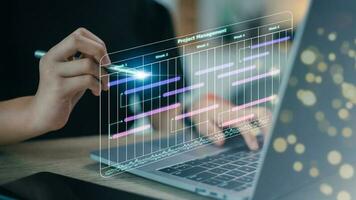 Business Project Management System. Project manager working on laptop and updating tasks and milestones progress planning with Gantt chart scheduling interface for the company on virtual screen. photo