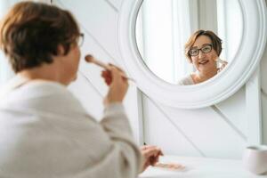 Smiling middle aged plus size woman doing makeup near mirror at home photo