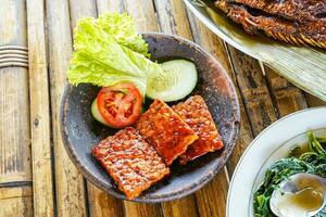 Grilled tempeh or fried tempeh with red barbecue sauce, vegetables and chili sauce served on an earthenware mortar photo