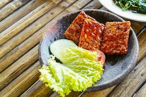 Grilled tempeh or fried tempeh with red barbecue sauce, vegetables and chili sauce served on an earthenware mortar photo