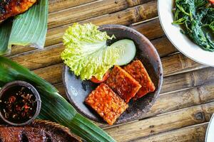 Grilled tempeh or fried tempeh with red barbecue sauce, vegetables and chili sauce served on an earthenware mortar photo