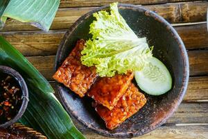 Grilled tempeh or fried tempeh with red barbecue sauce, vegetables and chili sauce served on an earthenware mortar photo