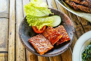 Grilled tempeh or fried tempeh with red barbecue sauce, vegetables and chili sauce served on an earthenware mortar photo