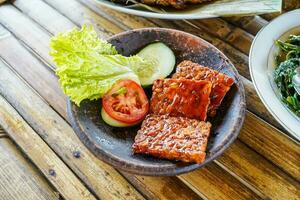 Grilled tempeh or fried tempeh with red barbecue sauce, vegetables and chili sauce served on an earthenware mortar photo