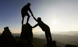 Silhouette of Two male  hikers climbing up mountain cliff and one of them giving helping hand. People helping and, team work concept. photo