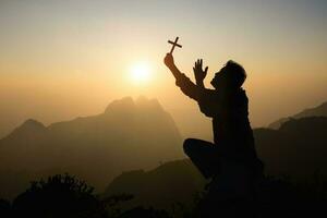 silueta de fiel hombre Orando con cristiano cruzar a puesta de sol como concepto para religión, fe, oración y espiritualidad. foto