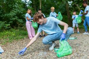 Hand of man picking up bottle into garbage bags while cleaning area in park. Volunteering, charity, people, ecology concept. Closeup volunteer collecting plastic trash in forest. World clean up m day photo