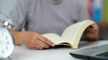 avvicinamento Immagine di asiatico donna seduta a un' tavolo lettura un' libro nel il vivente camera. video