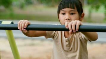 Little girl hanging on the horizontal bar on the playground. Cute young Asian girl hangs bar by hand to exercise at outdoor playground. video