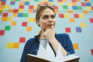 A student in a shirt and glasses holds a book in her hands on the street near the graffiti on the wall photo