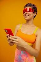 Woman blogger smiling in unusual red millennial glasses holding her phone and looking at the screen against an orange background in a stylish yellow top photo