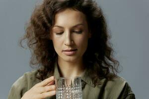 Focused serious adorable curly beautiful woman in casual khaki green shirt holds glass of water posing isolated on over gray blue background. Healthy lifestyle. Water is Life concept. Copy space photo