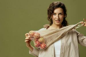 Smiling curly beautiful lady in linen casual shirt pulls string bag with fruit near chest looks at camera posing isolated on olive green background. Eco-friendly Natural products concept. Copy space photo