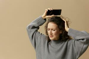 Crying curly beautiful female in gray casual sweater hold phone after hard breakup posing isolated on over beige pastel background. Social media, network, distance communication concept. Copy space photo